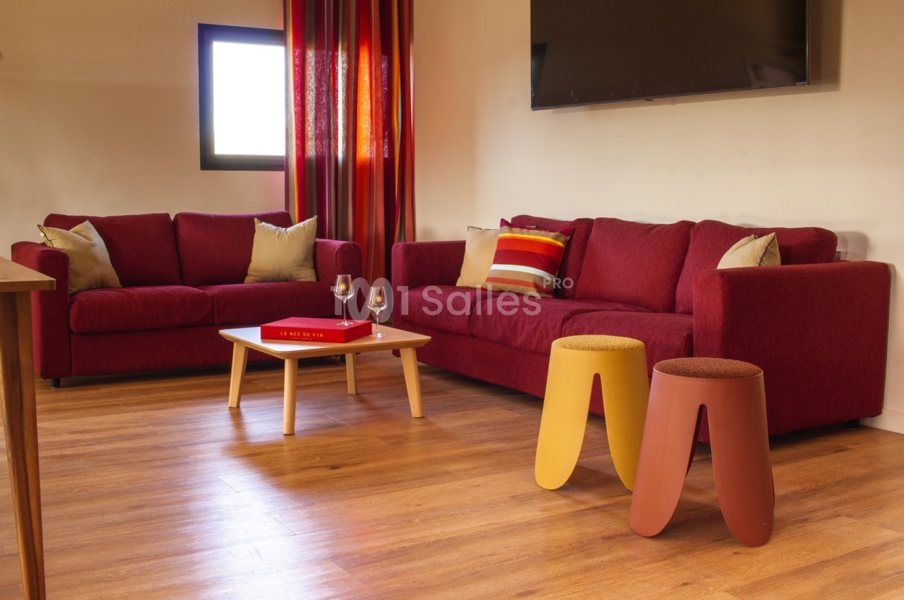 Salon avec deux canapés rouges, une table basse en bois, des tabourets colorés et un téléviseur fixé au mur.