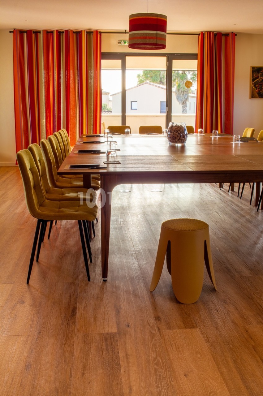 Salle de réunion lumineuse avec une grande table en bois, chaises jaunes, rideaux rouges et vue sur l'extérieur.