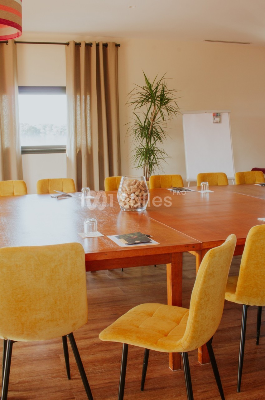 Salle de réunion lumineuse avec une grande table en bois, des chaises jaunes, un tableau blanc et une plante verte.