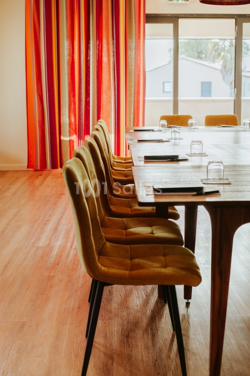 Salle de réunion avec table en bois, chaises jaunes alignées et rideaux rouges près de fenêtres.