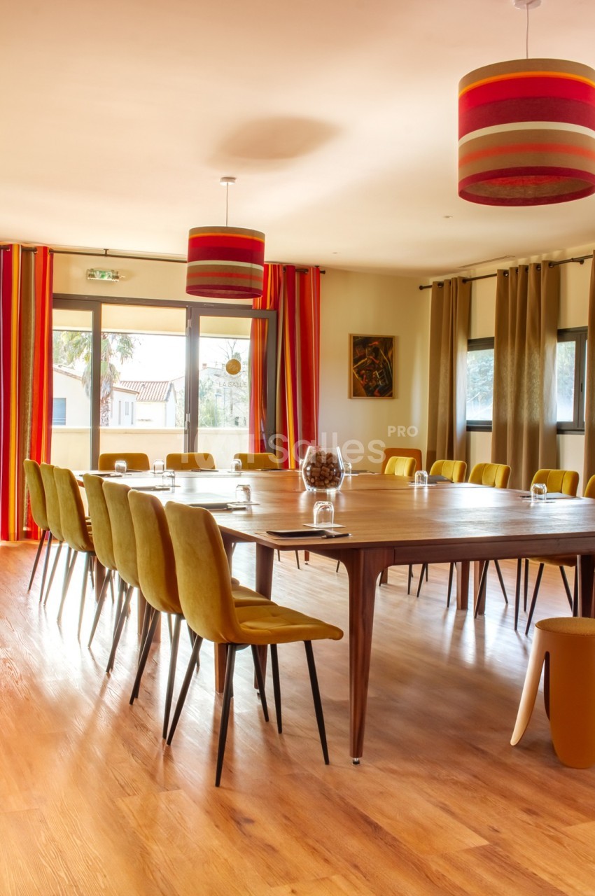 Salle de réunion lumineuse avec une grande table en bois entourée de chaises jaunes, rideaux colorés et suspensions…