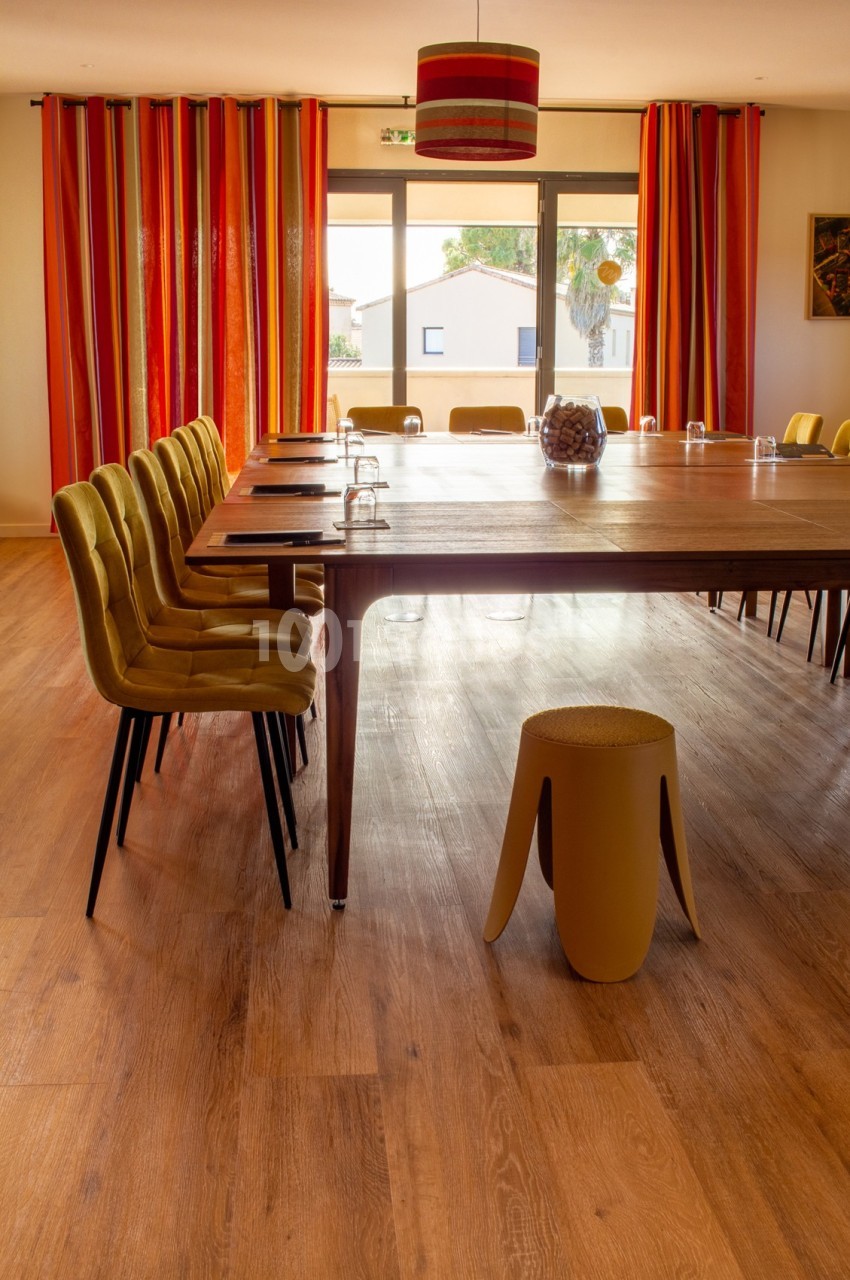 Salle de réunion lumineuse avec une grande table en bois, chaises jaunes, rideaux colorés et sol en parquet.