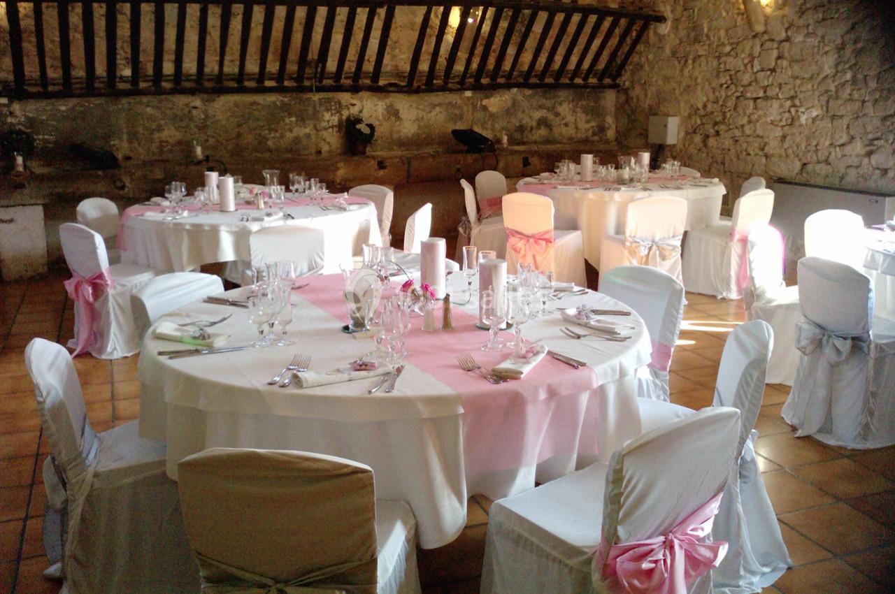 Salle de réception avec tables rondes dressées, nappes blanches et décorations roses, dans un cadre rustique en pierre.