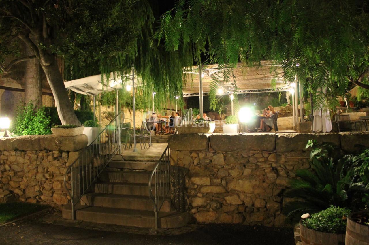 Terrasse extérieure éclairée la nuit, entourée de végétation et de murs en pierre, avec des tables occupées.