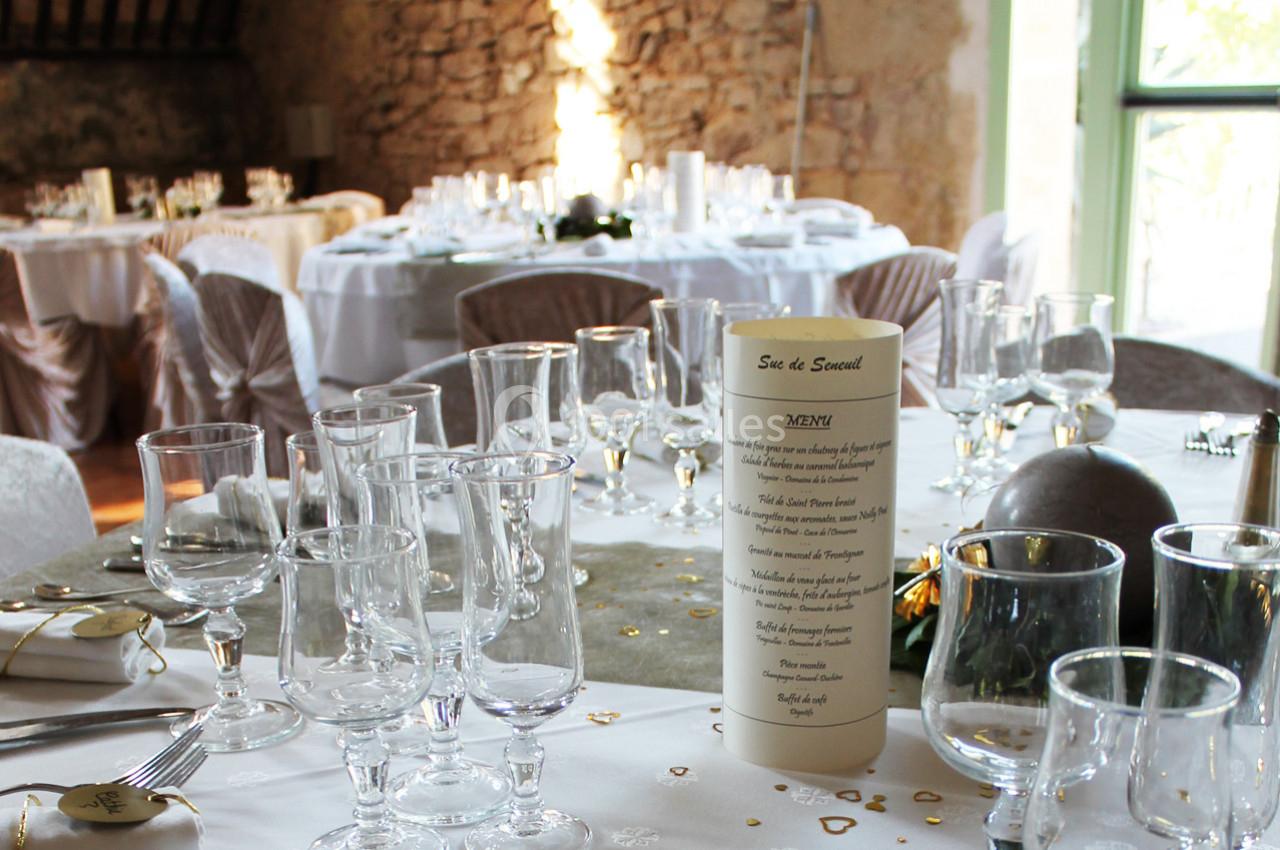 Salle de réception décorée avec des tables dressées, nappes blanches, menus et verres disposés pour un événement.