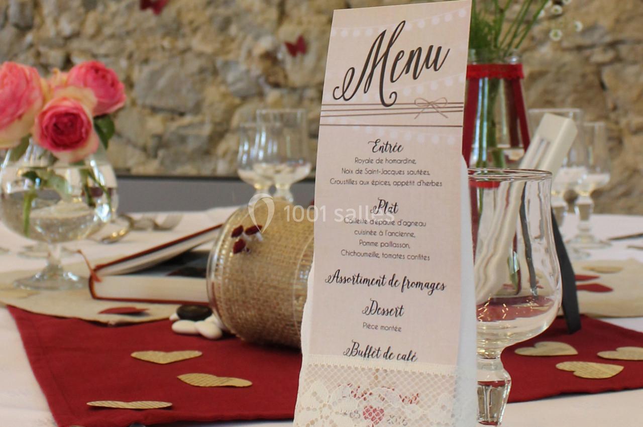 Menu de mariage posé sur une table décorée avec des fleurs, des pétales et des verres, devant un mur en pierre.