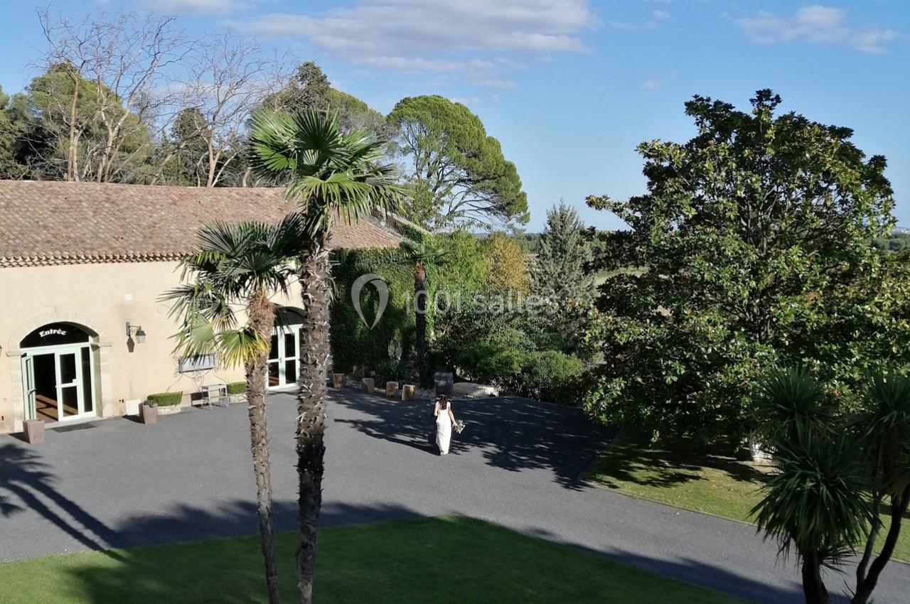 Vue d'un jardin avec pelouse, palmiers, bâtiment en pierre et une personne marchant sur une allée pavée.