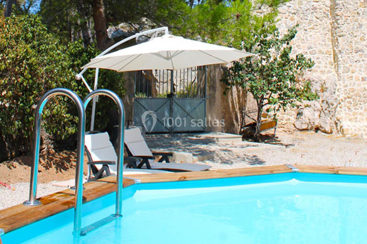 Piscine extérieure en bois avec parasol, transats et mur en pierre en arrière-plan, entourée de végétation.