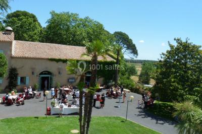Groupe de personnes rassemblées dans une cour extérieure verdoyante devant un bâtiment en pierre avec des volets bleus.