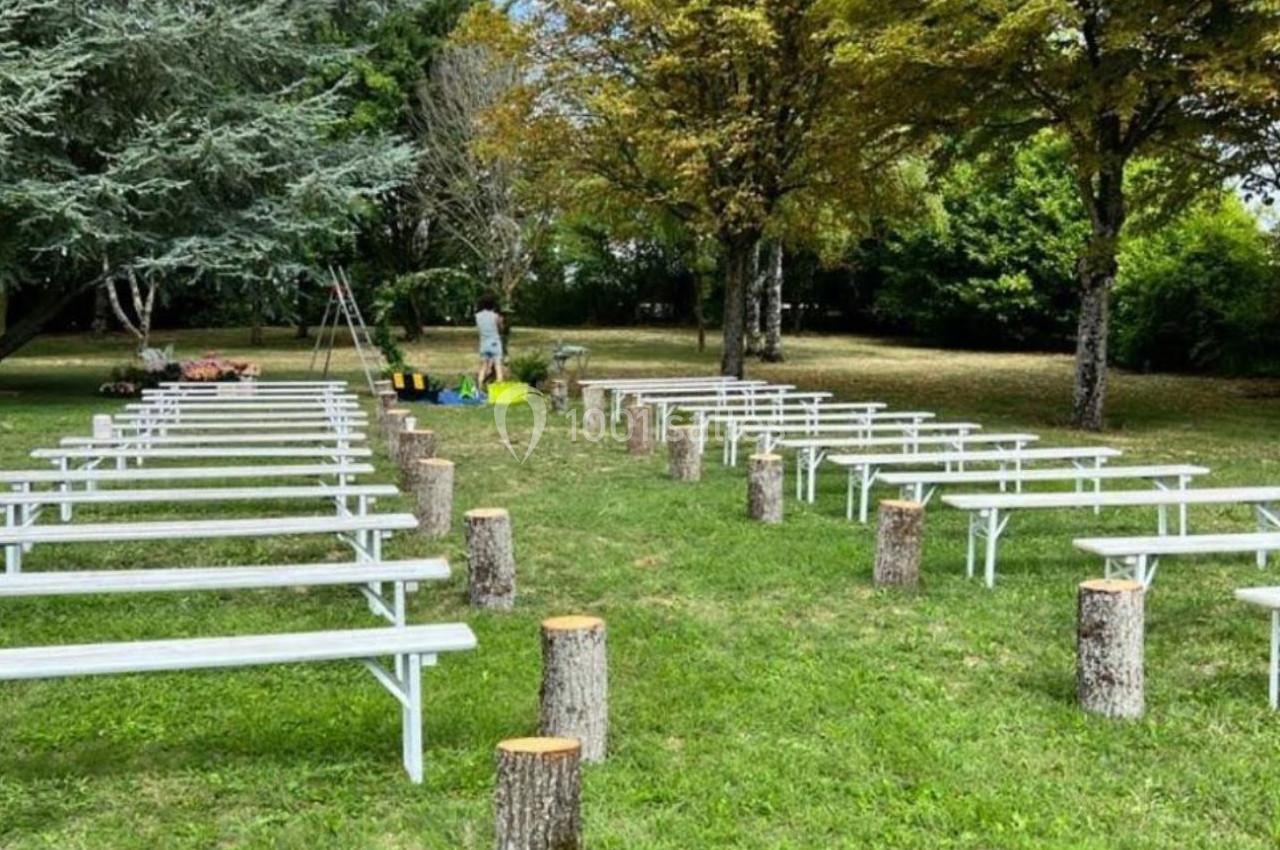 Location salle Villampuy (Eure-et-Loir) - Domaine de la Rainville #13 Bancs blancs alignés en extérieur sur une pelouse entourée d'arbres, avec des troncs d'arbres servant de décor.