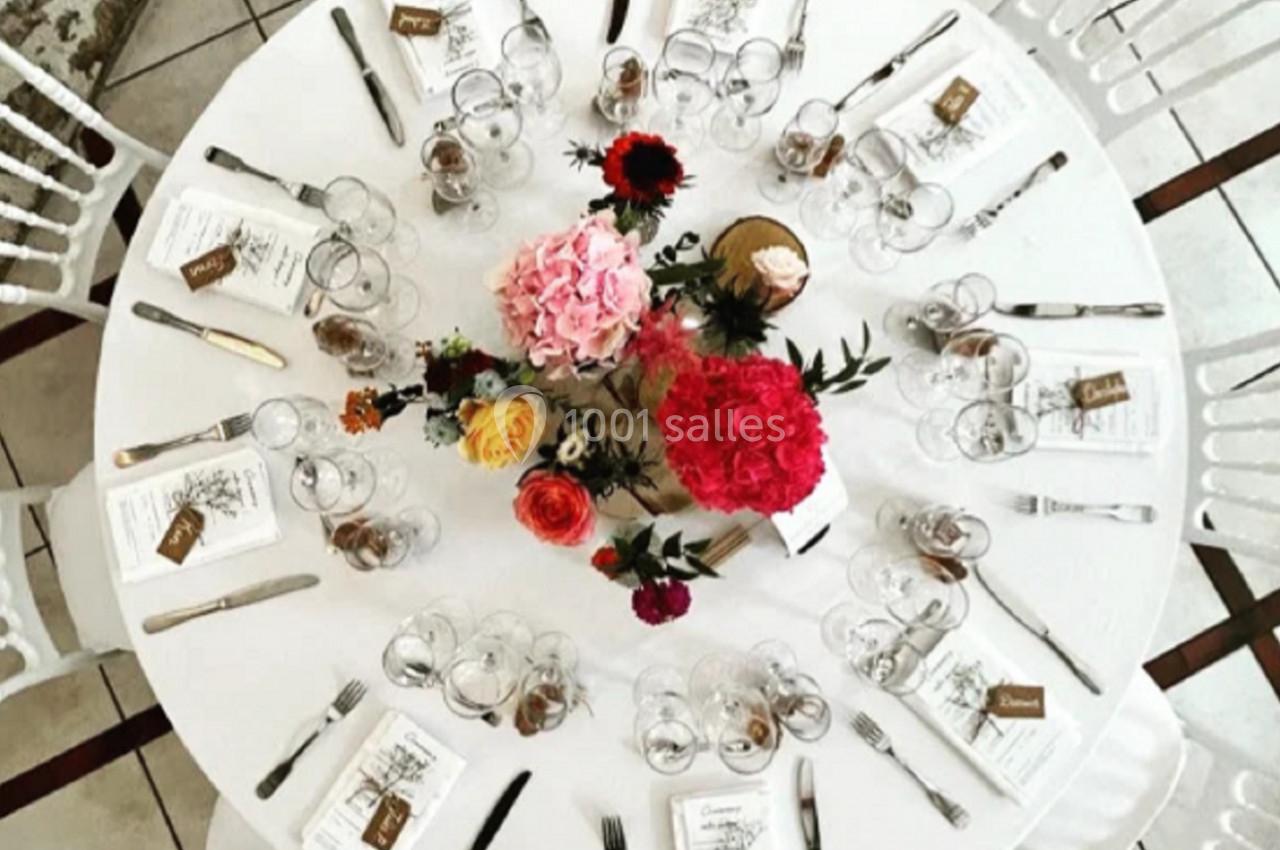 Location salle Villampuy (Eure-et-Loir) - Domaine de la Rainville #18 Table ronde dressée avec vaisselle, couverts, menus et centre de table floral coloré, entourée de chaises blanches.