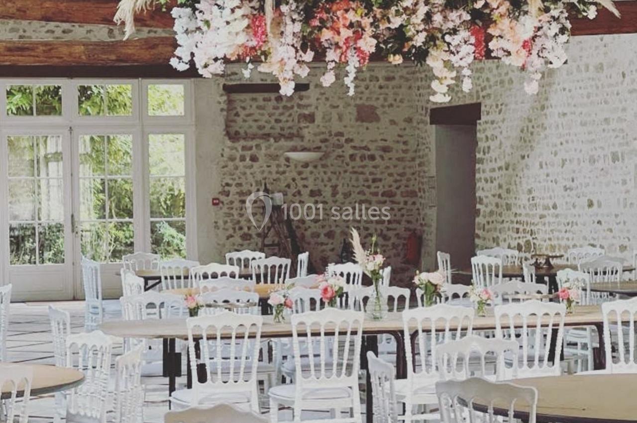 Location salle Villampuy (Eure-et-Loir) - Domaine de la Rainville #25 Salle de réception lumineuse avec tables rondes, chaises blanches et une décoration florale suspendue au plafond.