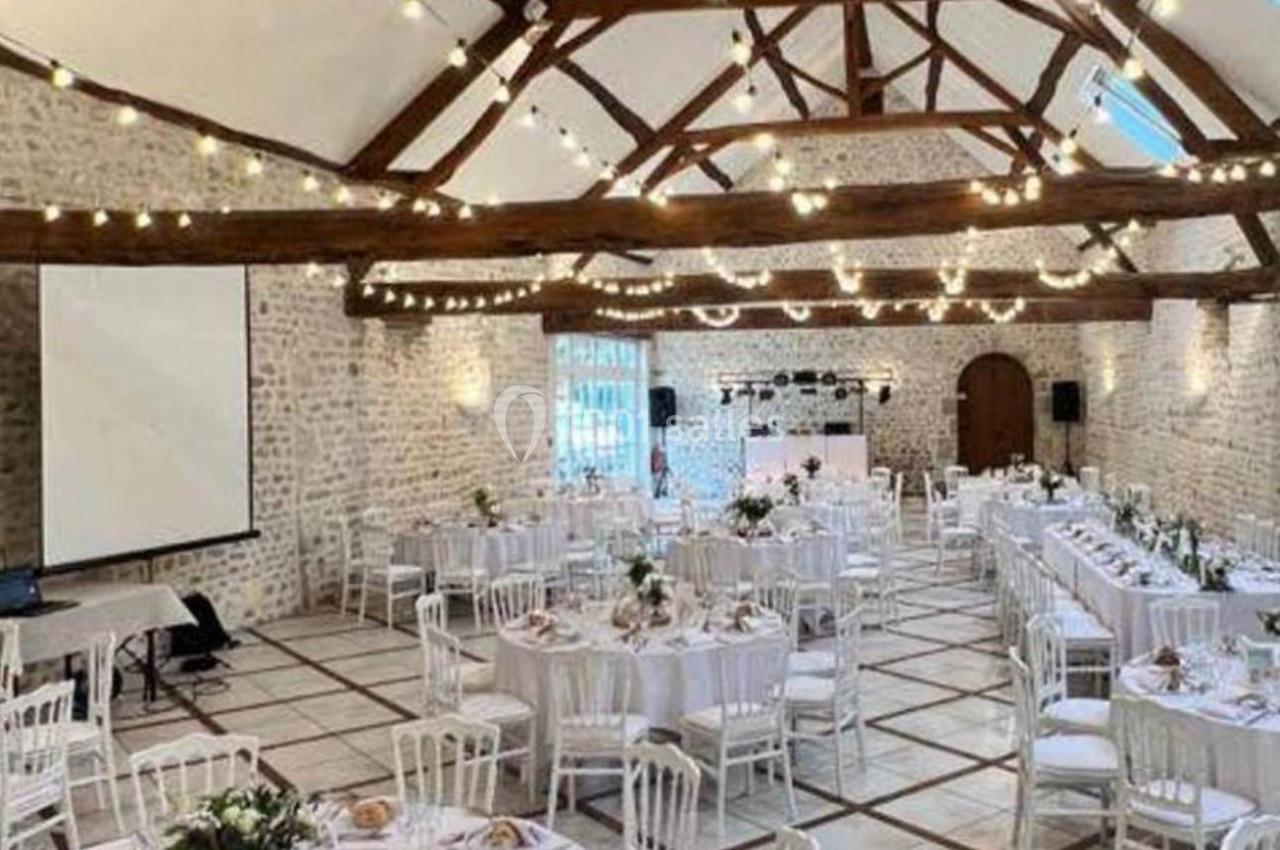 Location salle Villampuy (Eure-et-Loir) - Domaine de la Rainville #26 Salle de réception lumineuse avec poutres apparentes, tables rondes dressées et guirlandes lumineuses suspendues.
