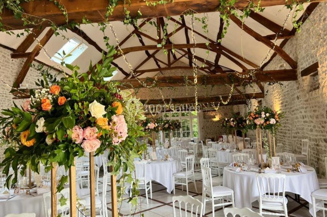 Salle de réception lumineuse avec poutres apparentes, tables décorées de nappes blanches et compositions florales colorées.