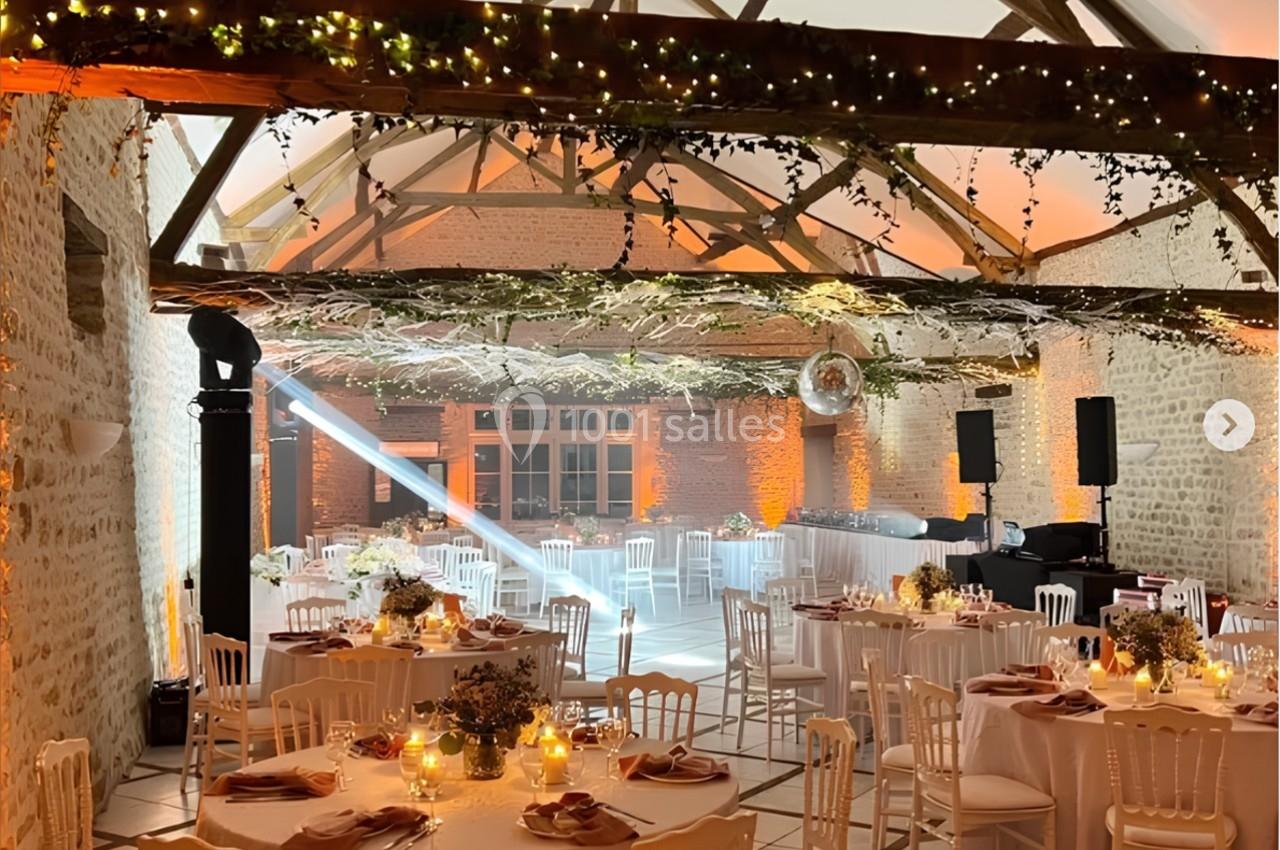 Location salle Villampuy (Eure-et-Loir) - Domaine de la Rainville #10 Salle de réception décorée avec des tables rondes, des chaises blanches, des lumières chaleureuses et des poutres apparentes.