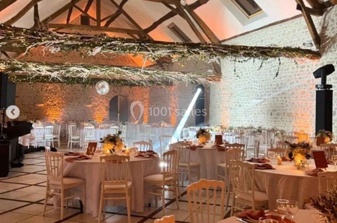 Location salle Villampuy (Eure-et-Loir) - Domaine de la Rainville #2 Salle de réception décorée avec des tables rondes, nappes blanches, guirlandes lumineuses et poutres apparentes.
