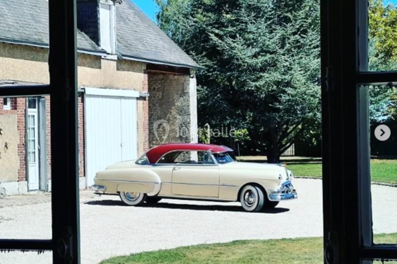 Location salle Villampuy (Eure-et-Loir) - Domaine de la Rainville #42 Voiture ancienne beige et rouge garée dans une cour gravillonnée, vue depuis l'intérieur d'une maison.