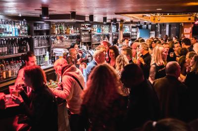 Salle avec un bar central, tabourets alignés, mezzanine avec tables, éclairage chaleureux et poutres apparentes.