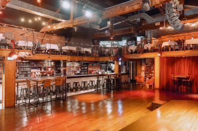 Salle avec un bar central, tabourets alignés, mezzanine avec tables, éclairage chaleureux et poutres apparentes.