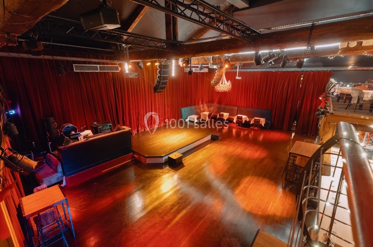 Salle de spectacle vide avec scène en bois, rideaux rouges, tables dressées et éclairage tamisé.