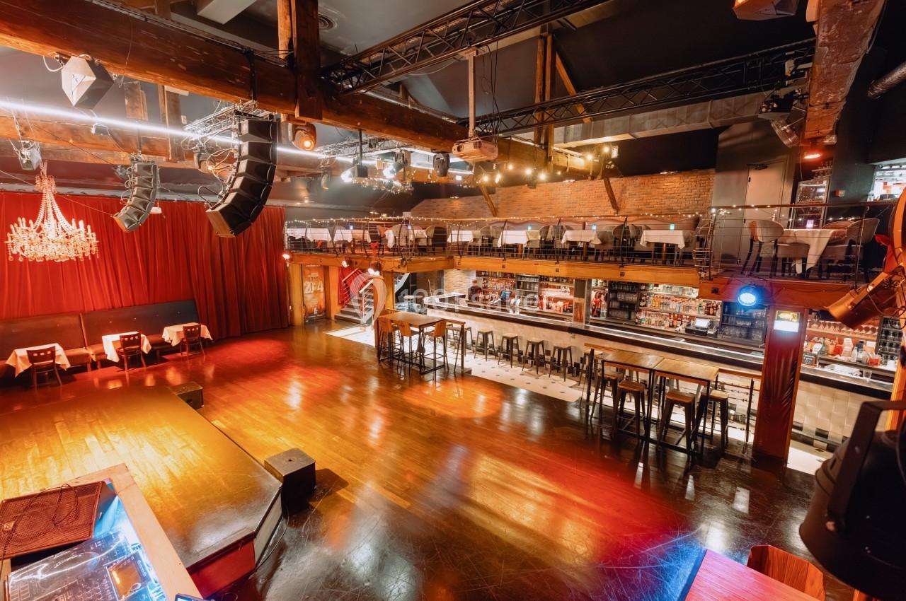 Salle de spectacle avec scène, tables, bar et mezzanine, éclairée par des lumières chaudes et un lustre.