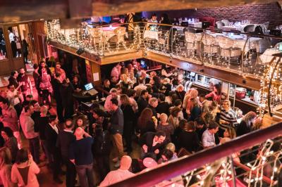 Salle avec un bar central, tabourets alignés, mezzanine avec tables, éclairage chaleureux et poutres apparentes.