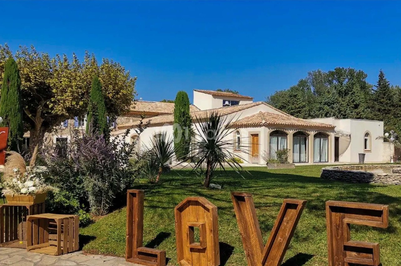Jardin avec pelouse, arbustes et sculptures en bois formant le mot ’LOVE’, devant une grande maison méditerranéenne.