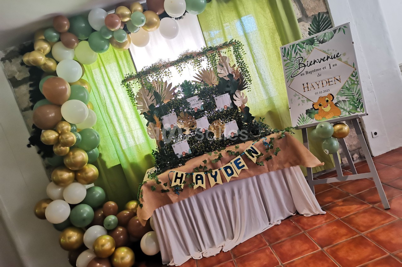 Décoration de fête avec ballons verts, blancs et dorés, table ornée de feuillages et panneau de bienvenue personnalisé.