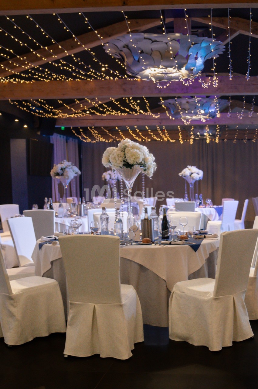 Salle de réception décorée pour un mariage avec tables rondes, nappes blanches, centres de table fleuris et guirlandes…