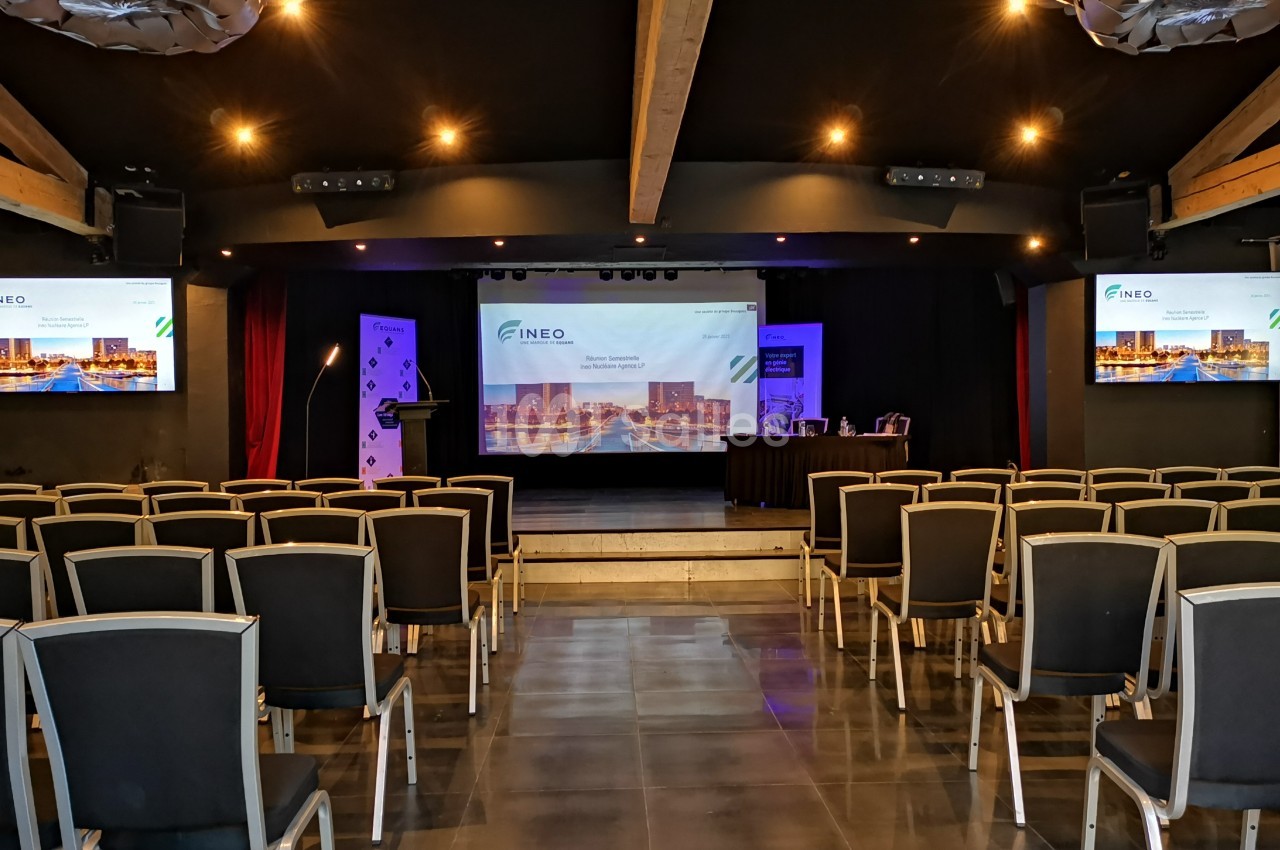 Salle de conférence vide avec des rangées de chaises, une scène équipée d'écrans et un projecteur allumé.