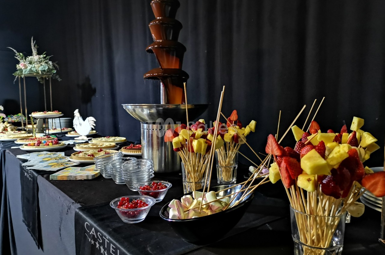 Fontaine de chocolat entourée de brochettes de fruits, desserts variés et décorations sur une table noire.