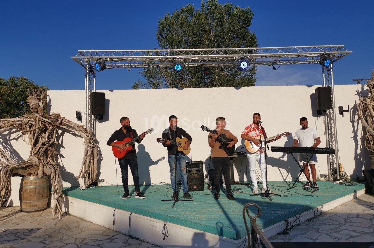 Un groupe de musiciens joue sur une petite scène extérieure sous un ciel bleu, entouré de décorations en bois.