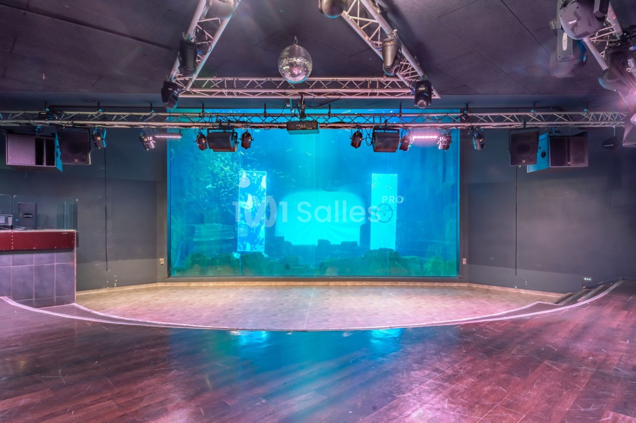 Scène vide avec un grand aquarium en arrière-plan, éclairée par des projecteurs suspendus au plafond.