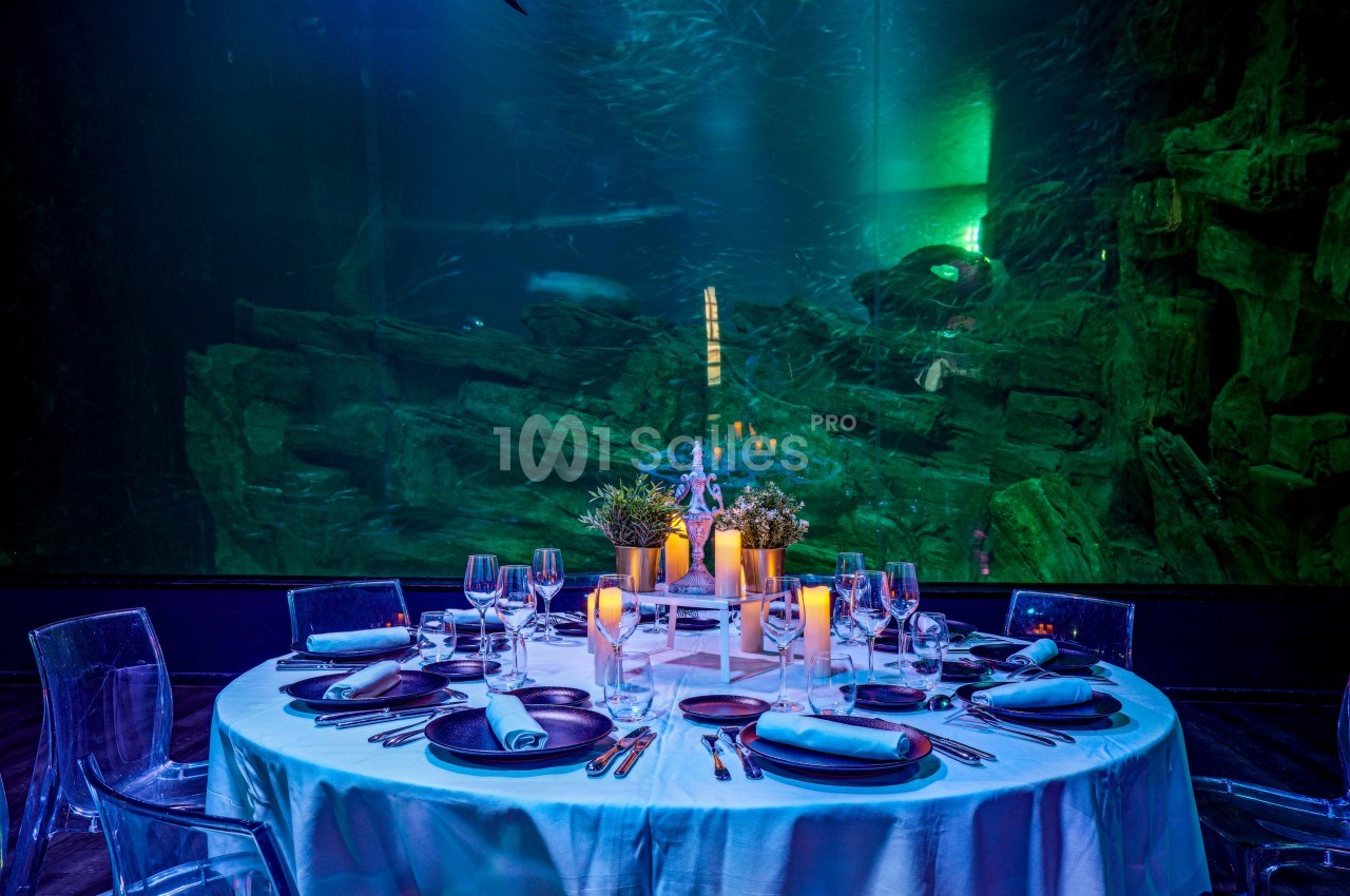 Table élégamment dressée avec des bougies et des plantes, placée devant un grand aquarium éclairé en bleu et vert.
