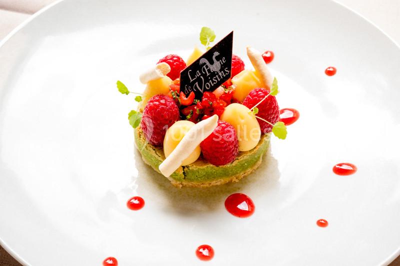 Tartelette garnie de framboises, crème pâtissière et décorations sur une assiette blanche avec coulis rouge.