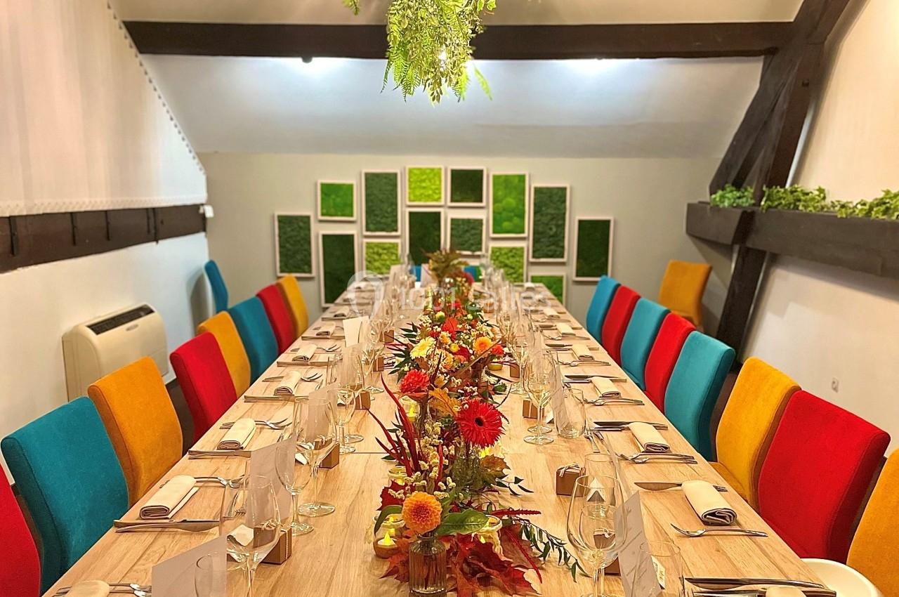 Table en bois dressée pour un repas, décorée de fleurs automnales, entourée de chaises colorées dans une salle lumineuse.