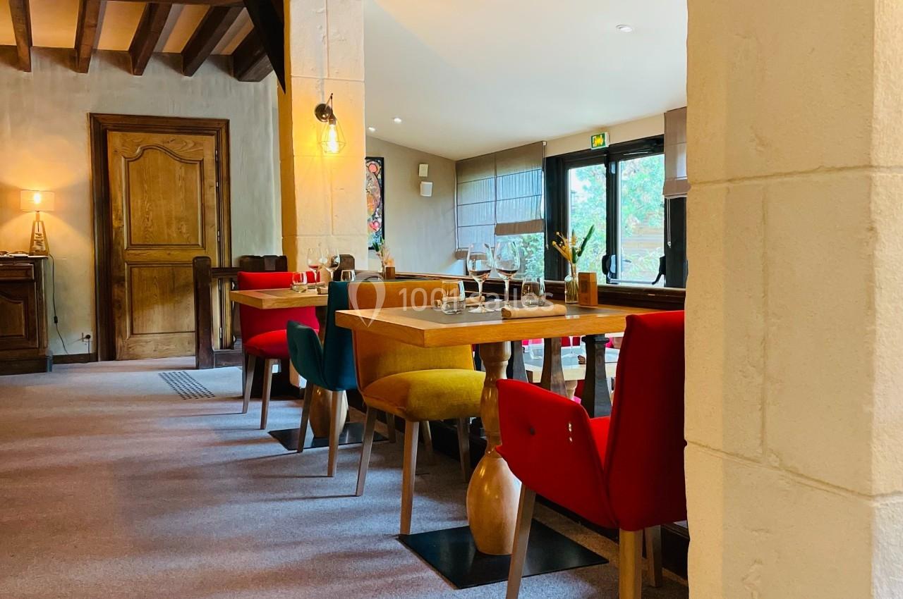 Salle à manger lumineuse avec tables en bois, chaises colorées et poutres apparentes, donnant sur un jardin.