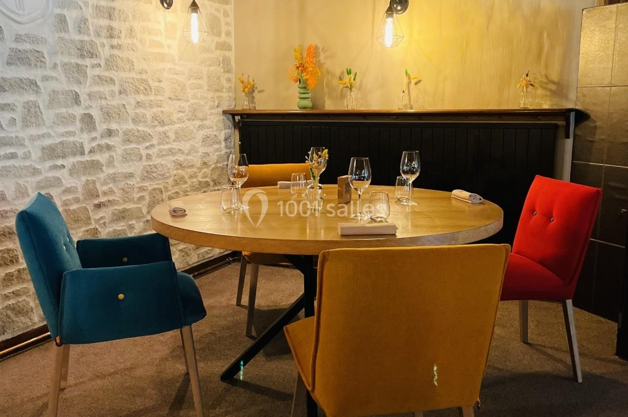 Salle à manger chaleureuse avec table ronde, chaises colorées, murs en pierre et éclairage suspendu.
