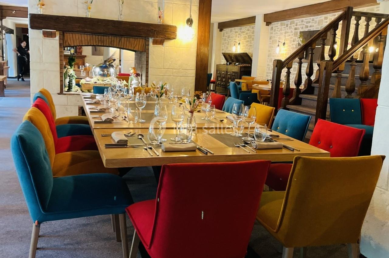 Salle à manger avec une grande table dressée, entourée de chaises colorées, sous un plafond en poutres apparentes.