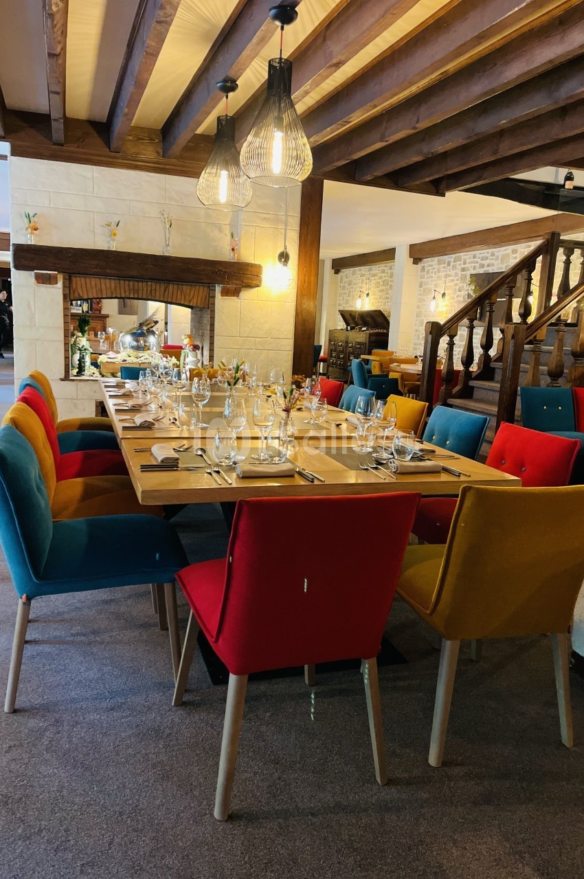 Salle à manger avec une grande table dressée, entourée de chaises colorées, sous un plafond en poutres apparentes.