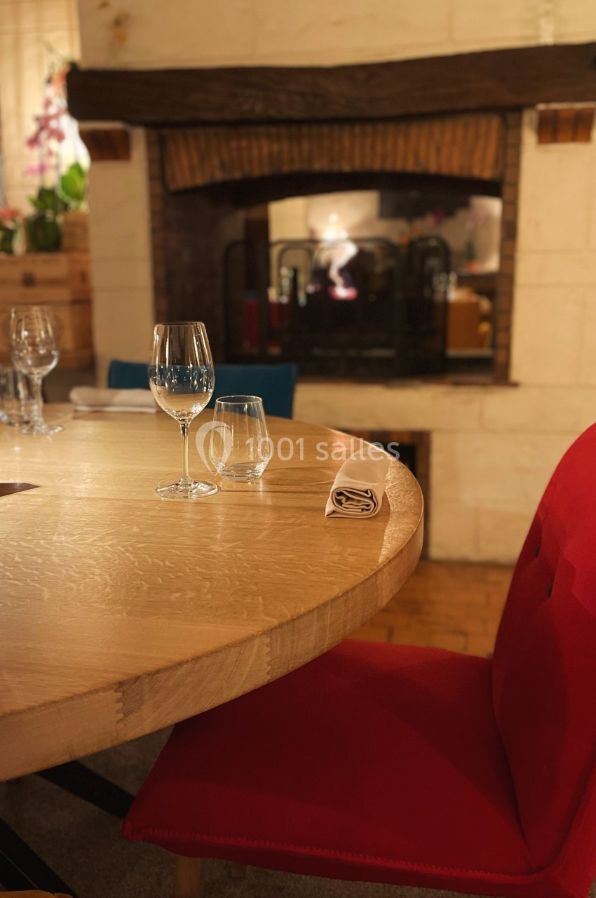 Table en bois dressée avec des verres, chaise rouge et cheminée allumée en arrière-plan dans un intérieur chaleureux.