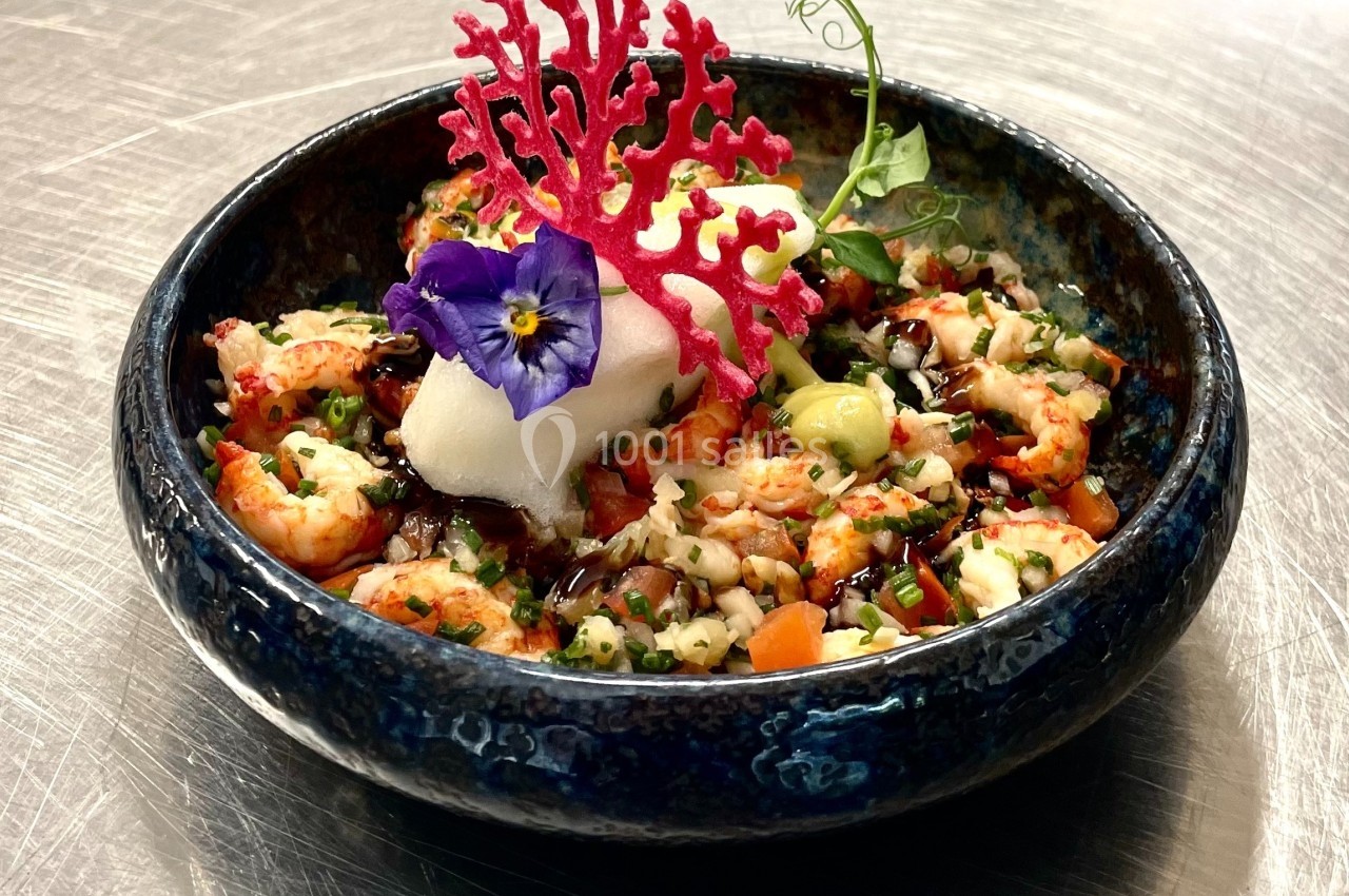 Plat raffiné dans un bol bleu, composé de langoustines, légumes, mousse blanche, fleur comestible et décoration rouge.