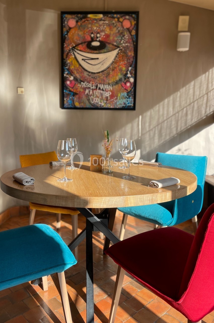 Table ronde dressée avec des verres et serviettes, entourée de chaises colorées, dans une salle lumineuse avec un tableau au…