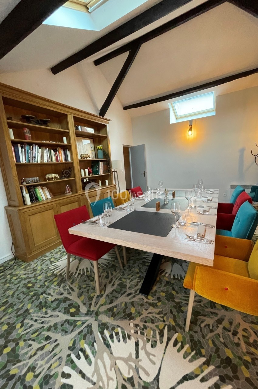 Salle à manger lumineuse avec table dressée, chaises colorées, bibliothèque en bois et poutres apparentes.