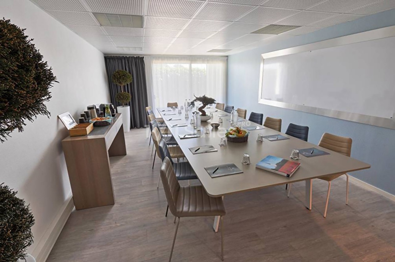 Salle de réunion lumineuse avec une grande table rectangulaire, des chaises, un tableau blanc et un buffet à boissons.