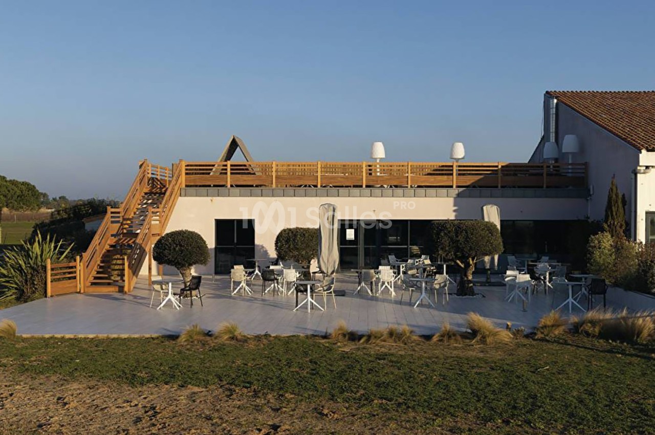 Terrasse extérieure d'un bâtiment avec tables, chaises, parasols et escaliers en bois, entourée de végétation.