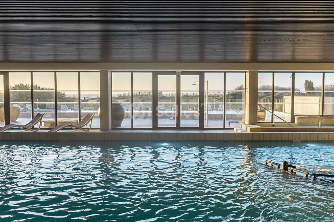Piscine intérieure avec grandes baies vitrées donnant sur une terrasse ensoleillée et un paysage extérieur.