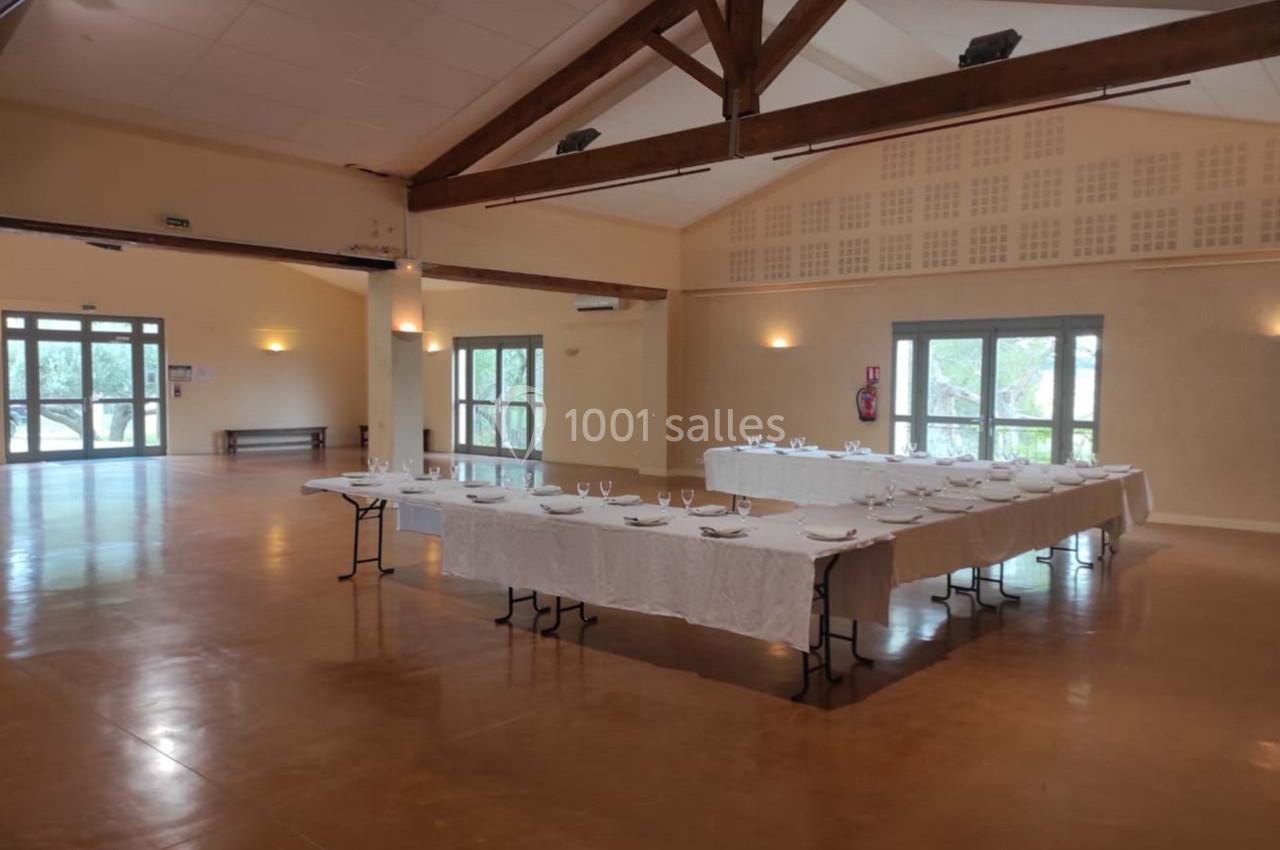 Salle de réception lumineuse avec tables dressées en U, nappes blanches et vaisselle, parquet et poutres apparentes.