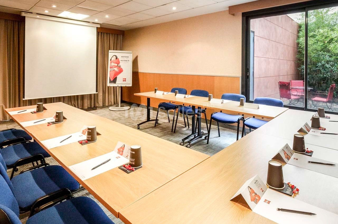 Salle de réunion lumineuse avec tables disposées en U, chaises bleues, écran de projection et vue sur un espace extérieur.