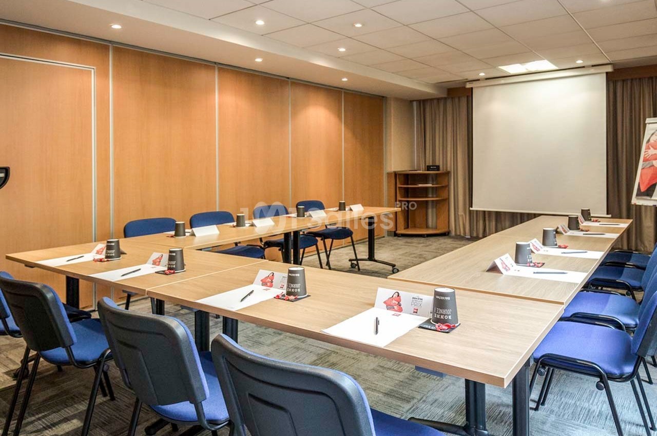 Salle de réunion lumineuse avec tables disposées en U, chaises bleues, écran de projection et fournitures de bureau.